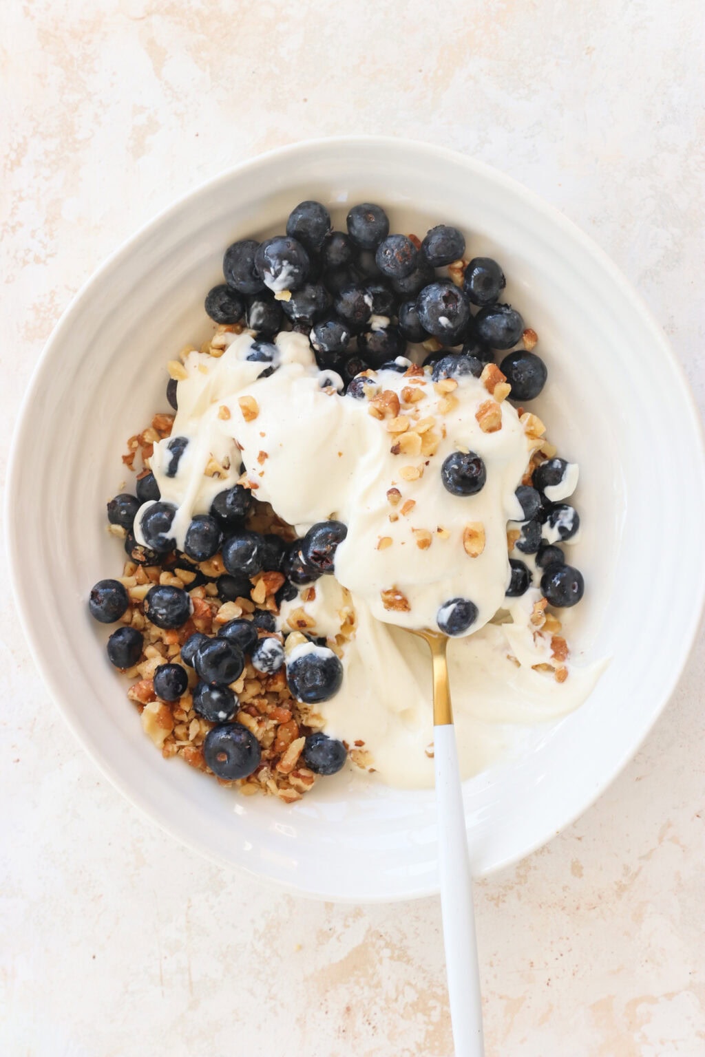 Ingredients for Frozen Blueberry Greek Yogurt Clusters added to a white bowl for mixing, including blueberries, walnuts, vanilla Greek yogurt, salt, chocolate chips, coconut oil