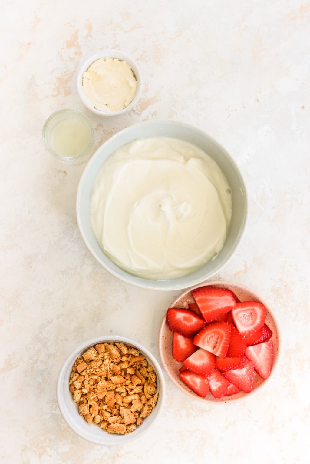 Ingredients for High Protein Frozen Greek Yogurt Cheesecake Bark with Strawberries in white bowls, including Greek yogurt, cream cheese, lemon, graham crackers, and strawberries